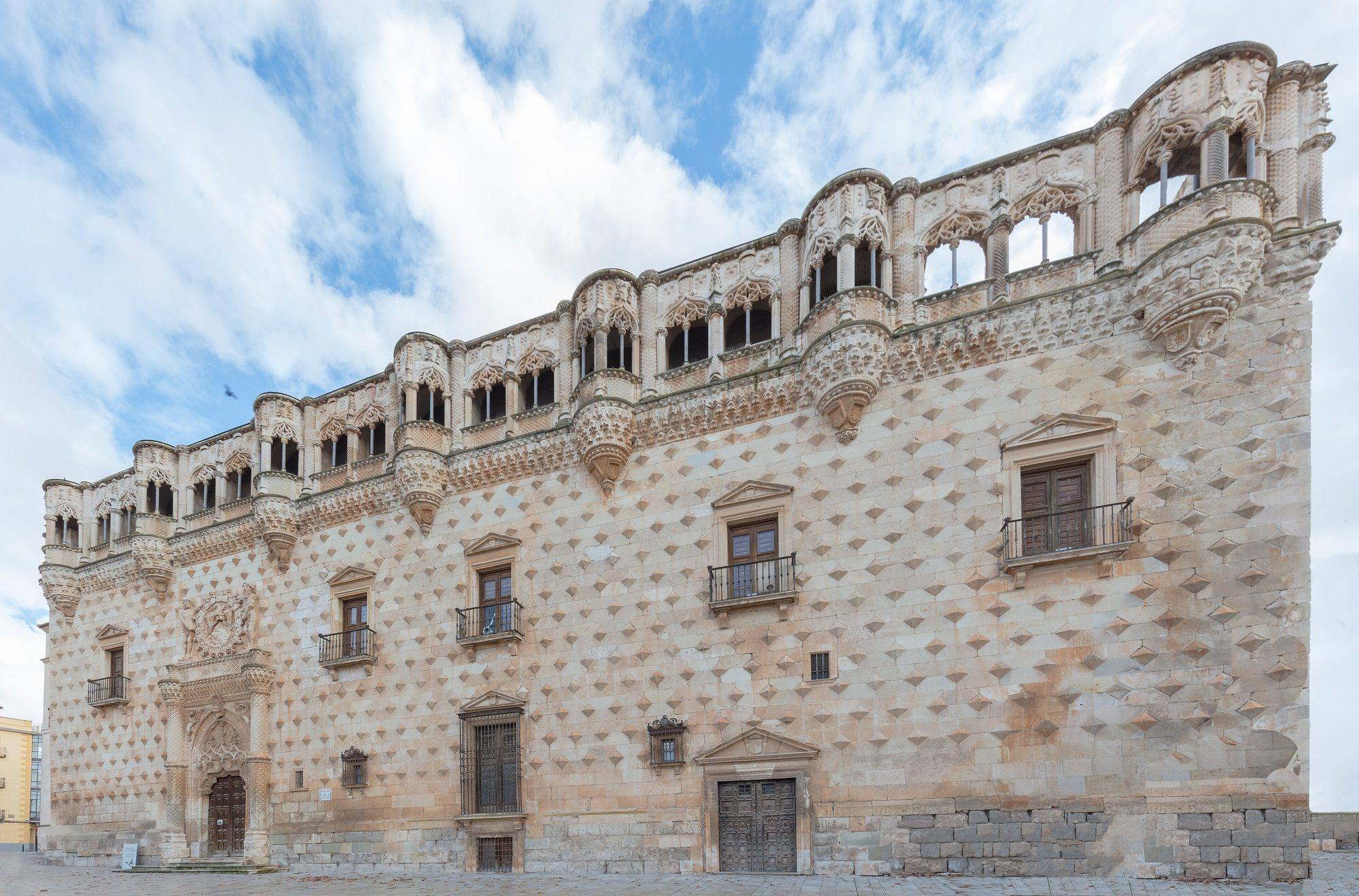 The main façade of the Palacio del Infantado in three-quarter view: a pale stone wall studded with rows of diamond-point bosses, crowned by a gallery of Gothic arches and corbelled turrets, with the carved main portal and coat of arms at left, under a blue sky with white clouds