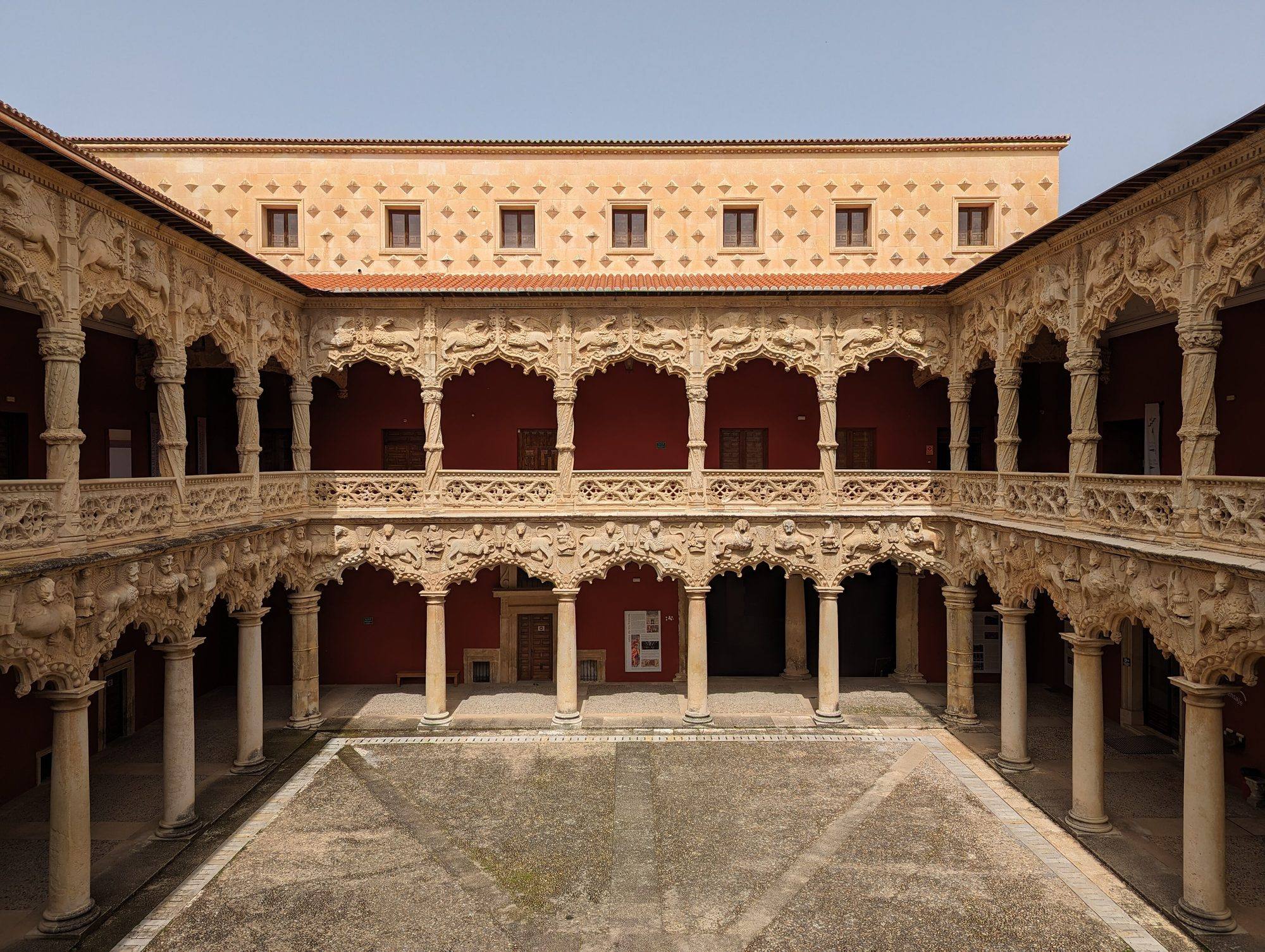 The Patio de los Leones of the Palacio del Infantado: two storeys of arcades with lobed arches carved with lions below and griffins above, the upper gallery on twisted columns, red walls behind, and a wall studded with diamond-point stones rising over the tiled roof