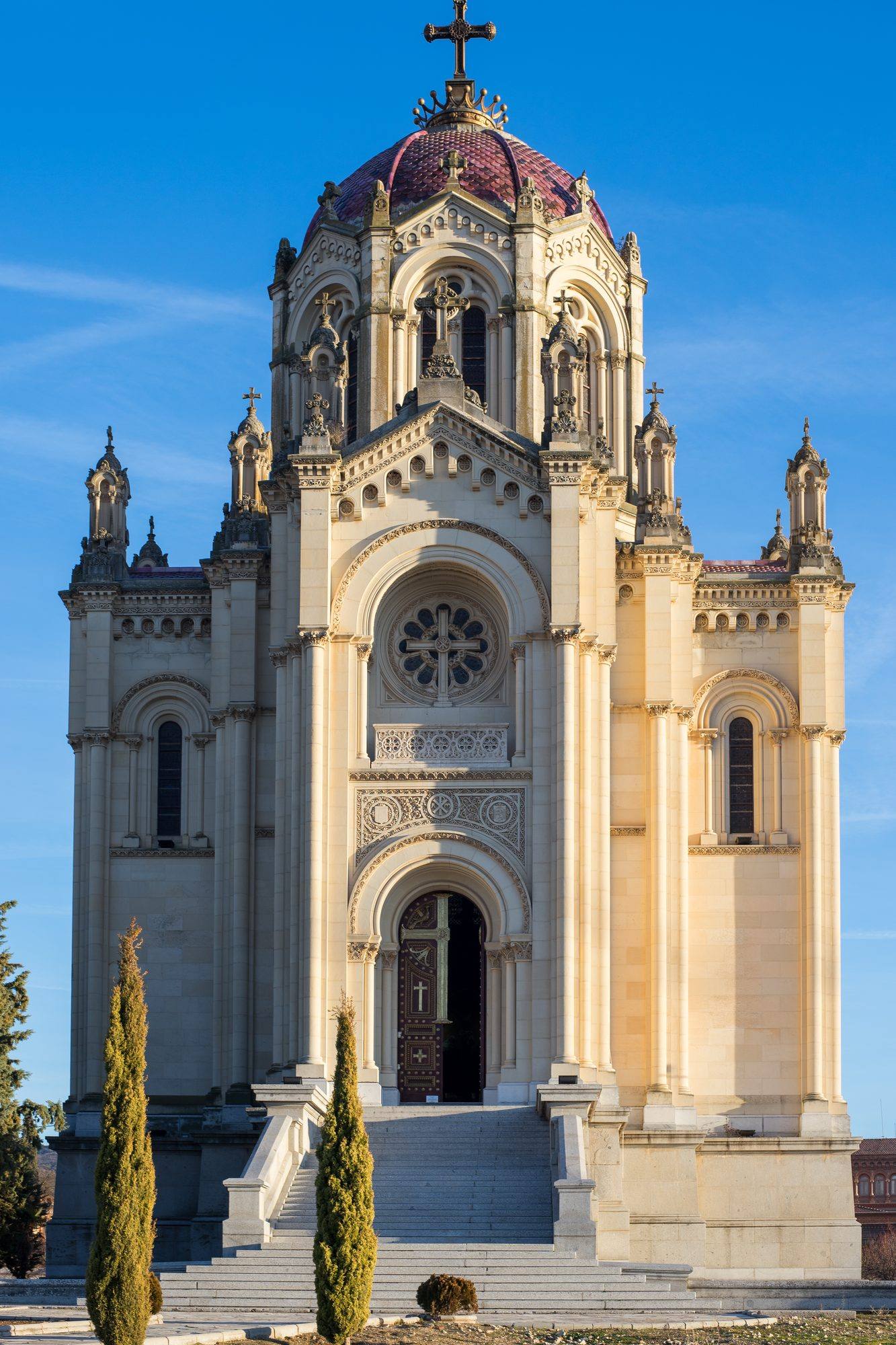 The Panteón de la Duquesa de Sevillano in low sunlight: a pale stone mausoleum with a rose window and an arched portal at the top of a wide staircase, crowned by a dome of glazed burgundy tiles and a cross, between slim cypresses under a deep blue sky