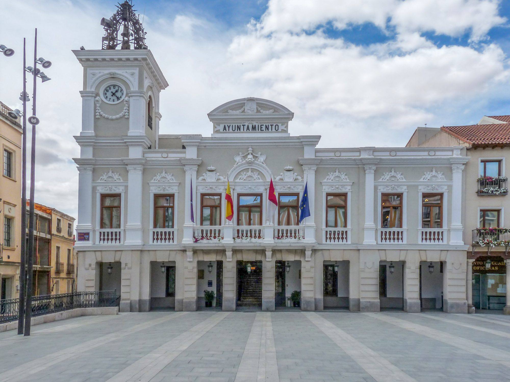 Guadalajara's town hall on the Plaza Mayor: a white early-20th-century façade with a clock tower topped by a wrought-iron bell frame, flags on the central balcony and an arcaded ground floor, facing an empty paved square under a cloudy blue sky