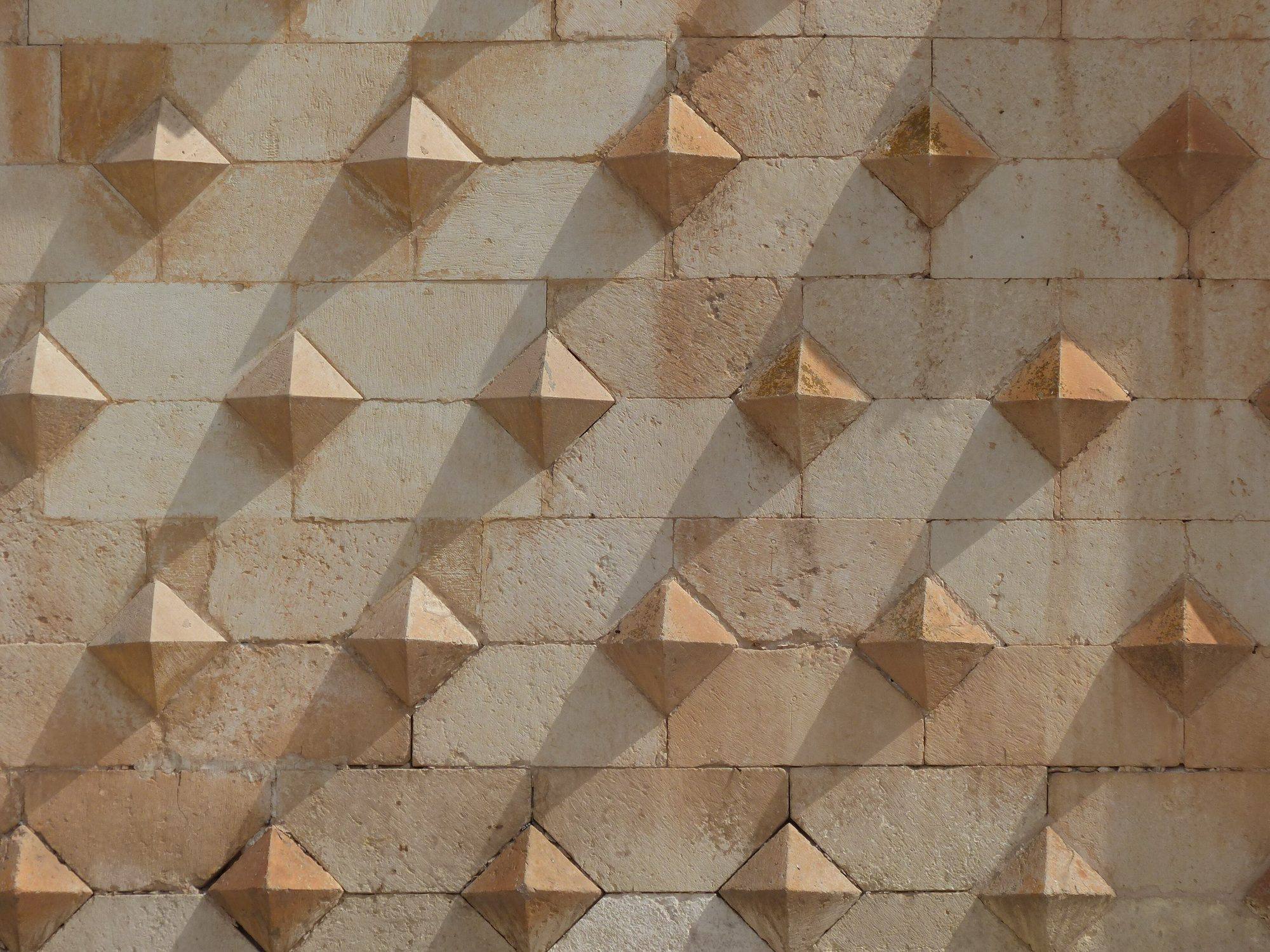 Close-up of the façade of the Palacio del Infantado: staggered rows of pyramid-shaped diamond-point stones projecting from the ashlar wall, casting long diagonal shadows in raking sunlight